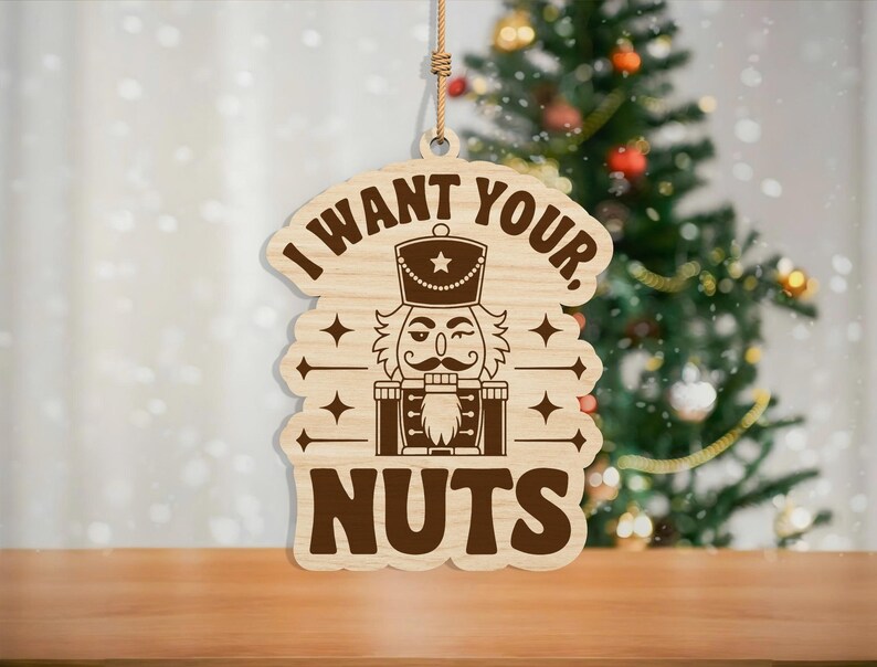 May include: Wooden Christmas ornament with the text "I WANT YOUR, NUTS" and a nutcracker design. The ornament is brown and hangs from a natural twine string. A Christmas tree is visible in the background.