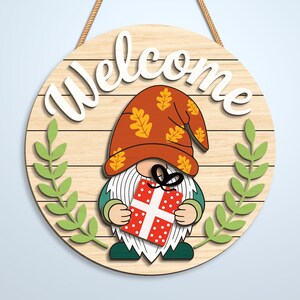 K&ouml;nnte beinhalten: Ein rundes Holzschild mit heller Holzmaserung und dem wei&szlig;en Schriftzug Welcome oben. Ein Gnom mit einem rostfarbenen Hut, der mit gelben Eichenbl&auml;ttern verziert ist, h&auml;lt ein rotes Geschenk mit wei&szlig;en Punkten und einer schwarzen Schleife.