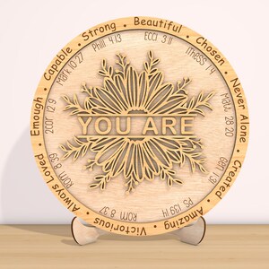 May include: A round wooden plaque with a floral design and the words "YOU ARE" in the center. The plaque is surrounded by a circle of words and bible verses, including "Strong", "Beautiful", "Chosen", "Never Alone", "Created", "Amazing", "Victorious", "Always Loved", "Enough", "Capable", and "Matt. 28:20".
