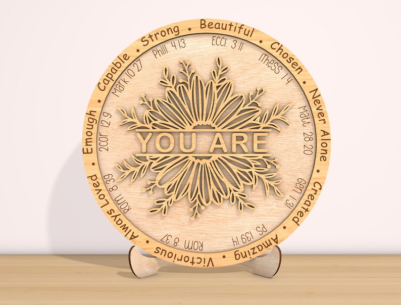 May include: A round wooden plaque with a floral design and the words "YOU ARE" in the center. The plaque is surrounded by a circle of words and bible verses, including "Strong", "Beautiful", "Chosen", "Never Alone", "Created", "Amazing", "Victorious", "Always Loved", "Enough", "Capable", and "Matt. 28:20".