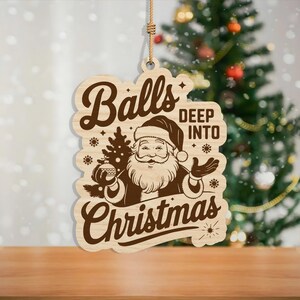 May include: Wooden Christmas ornament with a Santa Claus graphic and the words "Balls Deep Into Christmas". The ornament is light brown with a dark brown graphic and a string for hanging. A Christmas tree is in the background.