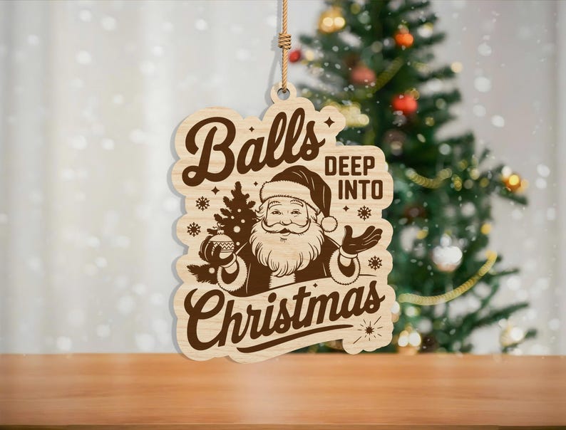 May include: Wooden Christmas ornament with a Santa Claus graphic and the words "Balls Deep Into Christmas". The ornament is light brown with a dark brown graphic and a string for hanging. A Christmas tree is in the background.