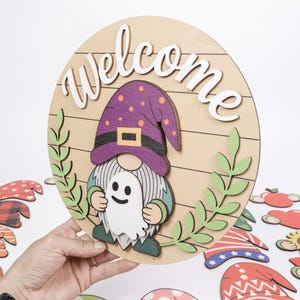 K&ouml;nnte beinhalten: Ein rundes Holzschild mit dem Wort Welcome in wei&szlig;er Schreibschrift, einem Zwerg mit violettem Hut mit orangefarbenen Punkten und einer wei&szlig;en Geisterfigur. Das Design umfasst gr&uuml;ne Blattakzente auf hellem Holz mit horizontalen Linien.