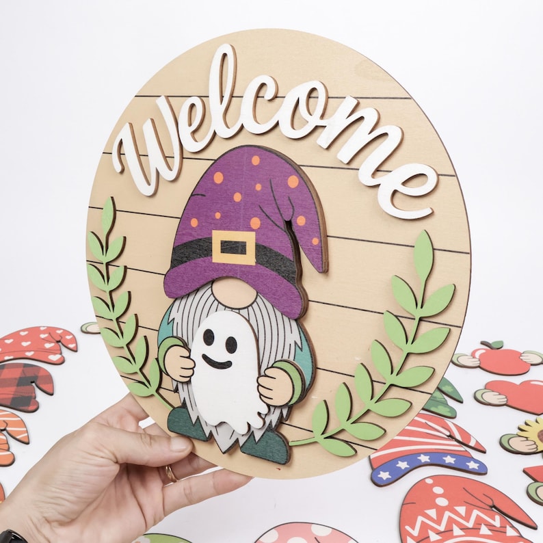 K&ouml;nnte beinhalten: Ein rundes Holzschild mit dem Wort Welcome in wei&szlig;er Schreibschrift, einem Zwerg mit violettem Hut mit orangefarbenen Punkten und einer wei&szlig;en Geisterfigur. Das Design umfasst gr&uuml;ne Blattakzente auf hellem Holz mit horizontalen Linien.
