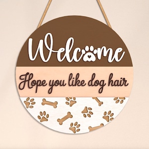 May include: Round wooden welcome sign with a brown and beige color scheme. The sign features the word "Welcome" in white script, a paw print, and the phrase "Hope you like dog hair." Paw prints and bones decorate the bottom.