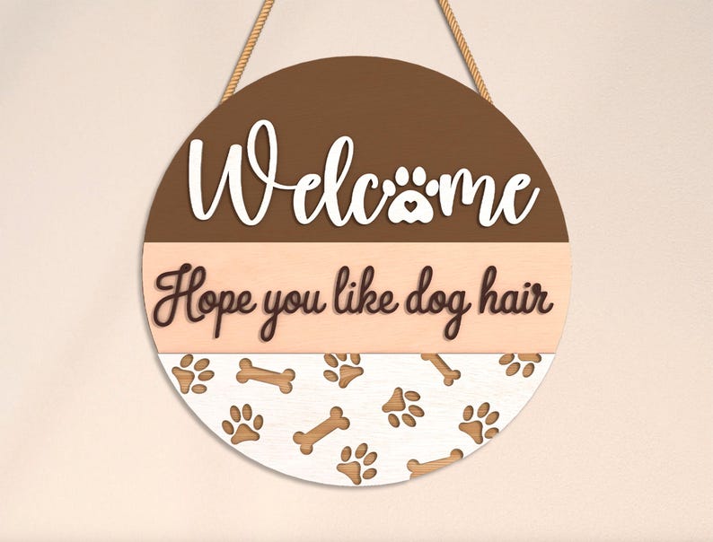 May include: Round wooden welcome sign with a brown and beige color scheme. The sign features the word "Welcome" in white script, a paw print, and the phrase "Hope you like dog hair." Paw prints and bones decorate the bottom.