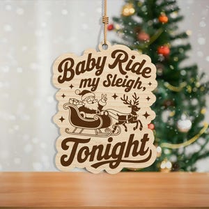 May include: Wooden Christmas ornament with the text "Baby Ride my Sleigh, Tonight". The ornament features a Santa Claus in a sleigh pulled by reindeer. The ornament is hanging in front of a Christmas tree.