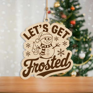 May include: Wooden Christmas ornament with the phrase "LET'S GET Frosted" and a snowman graphic. The snowman wears a hat and scarf, holding a bottle. The ornament hangs from a jute string, with a Christmas tree in the background.