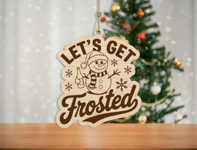 May include: Wooden Christmas ornament with the phrase "LET'S GET Frosted" and a snowman graphic. The snowman wears a hat and scarf, holding a bottle. The ornament hangs from a jute string, with a Christmas tree in the background.