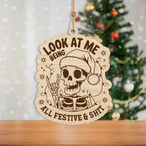 May include: Wooden Christmas ornament featuring a skeleton wearing a Santa hat and string lights. The ornament has the text "LOOK AT ME BEING ALL FESTIVE & SHIT" engraved on it. The ornament is hanging by a natural fiber string.