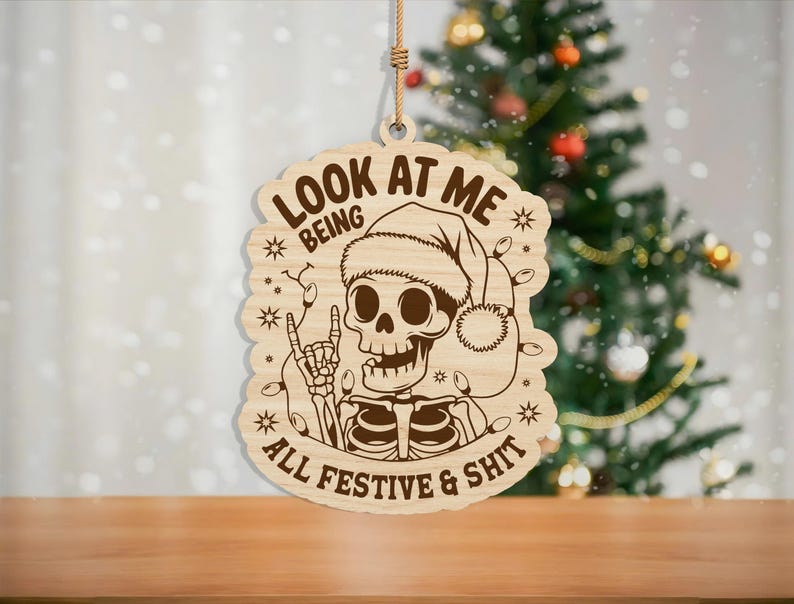 May include: Wooden Christmas ornament featuring a skeleton wearing a Santa hat and string lights. The ornament has the text "LOOK AT ME BEING ALL FESTIVE & SHIT" engraved on it. The ornament is hanging by a natural fiber string.