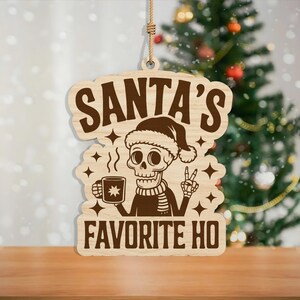 May include: Wooden Christmas ornament with the text "SANTA'S FAVORITE HO". The ornament features a skeleton wearing a Santa hat and scarf, holding a mug and giving a peace sign. The ornament is hanging by a rope.