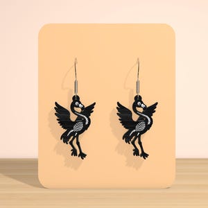 May include: A pair of black bird-shaped earrings with a skeleton design. The earrings have a silver hook and a spring detail. The birds have outstretched wings and are displayed on a peach-colored background.
