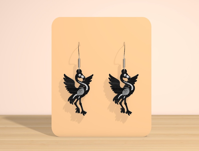 May include: A pair of black bird-shaped earrings with a skeleton design. The earrings have a silver hook and a spring detail. The birds have outstretched wings and are displayed on a peach-colored background.