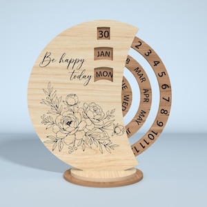 Puede incluir: Calendario perpetuo de madera con dise&ntilde;o floral y la frase "Be happy today". El calendario muestra la fecha, el mes y el d&iacute;a. Tiene una base redonda y anillos giratorios para el mes y el d&iacute;a.