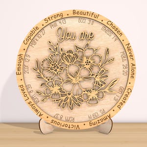 May include: A round wooden plaque with a floral design and the words "You are" in the center. The plaque is surrounded by a circle of words and bible verses, including "Strong", "Beautiful", "Chosen", "Never Alone", "Created", "Amazing", "Victorious", "Always Loved", "Enough", "Capable".