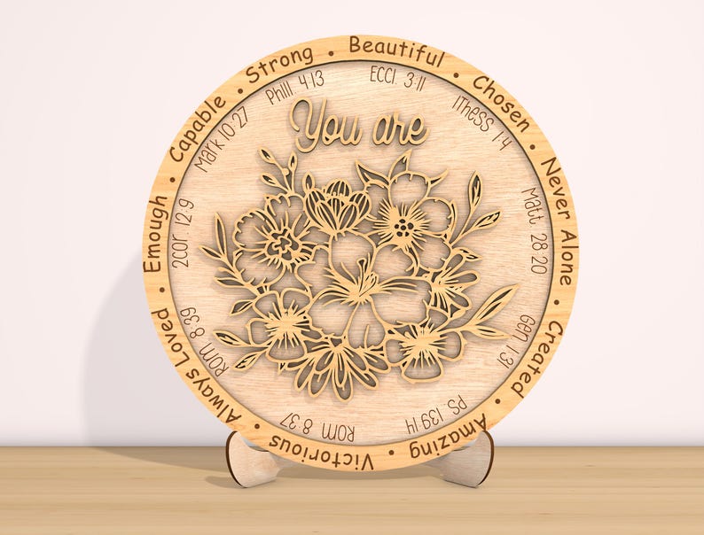 May include: A round wooden plaque with a floral design and the words "You are" in the center. The plaque is surrounded by a circle of words and bible verses, including "Strong", "Beautiful", "Chosen", "Never Alone", "Created", "Amazing", "Victorious", "Always Loved", "Enough", "Capable".