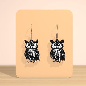 May include: A pair of black owl earrings with a skeleton design. The earrings are displayed on a peach-colored background. The owl earrings have a detailed skeleton design and are suspended from silver-colored hooks.