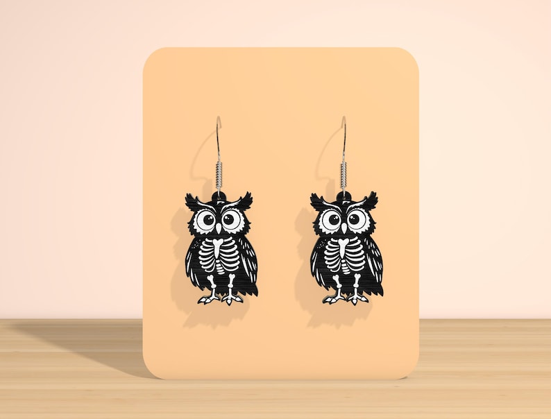 May include: A pair of black owl earrings with a skeleton design. The earrings are displayed on a peach-colored background. The owl earrings have a detailed skeleton design and are suspended from silver-colored hooks.