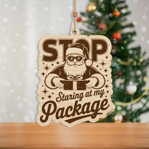 May include: Wooden Christmas ornament featuring a Santa Claus graphic wearing sunglasses and holding a gift. The ornament has the text "STOP Staring at my Package" in a playful font. The ornament hangs from a natural twine.