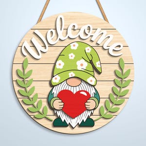 K&ouml;nnte beinhalten: Ein rundes Holzschild mit heller Holzmaserung und dem wei&szlig;en Schriftzug Welcome oben. Ein Zwerg mit gr&uuml;nem Blumenhut und wei&szlig;em Bart h&auml;lt ein gro&szlig;es rotes Herz. Zwei gr&uuml;ne Blattzweige rahmen den Zwerg auf beiden Seiten des Schildes ein.