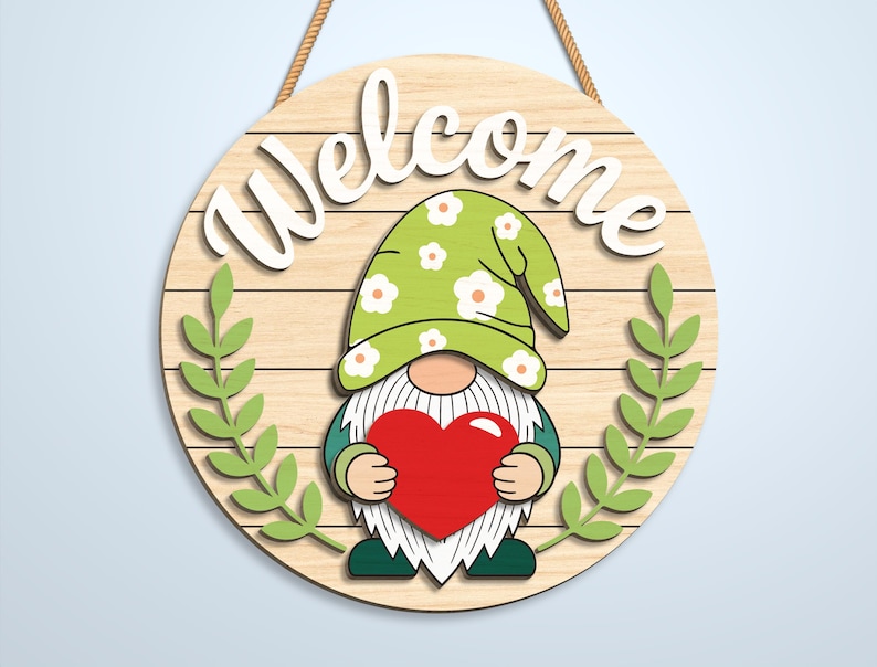 K&ouml;nnte beinhalten: Ein rundes Holzschild mit heller Holzmaserung und dem wei&szlig;en Schriftzug Welcome oben. Ein Zwerg mit gr&uuml;nem Blumenhut und wei&szlig;em Bart h&auml;lt ein gro&szlig;es rotes Herz. Zwei gr&uuml;ne Blattzweige rahmen den Zwerg auf beiden Seiten des Schildes ein.