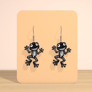May include: A pair of black frog-shaped earrings with white skeleton detailing. The earrings have a smiling face and are suspended from silver-colored hooks. The earrings are displayed on a peach-colored background.