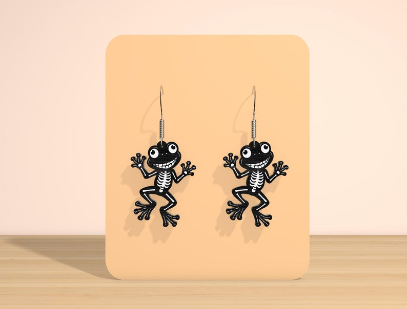May include: A pair of black frog-shaped earrings with white skeleton detailing. The earrings have a smiling face and are suspended from silver-colored hooks. The earrings are displayed on a peach-colored background.