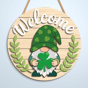 K&ouml;nnte beinhalten: Ein rundes h&auml;ngendes Holzschild mit dem Wort Welcome in wei&szlig;er Schreibschrift. In der Mitte befindet sich ein Gnom mit einem gr&uuml;nen Hut mit Kleeblattmuster, der ein gro&szlig;es gr&uuml;nes Kleeblatt h&auml;lt. Zwei hellgr&uuml;ne Blattzweige umrahmen den Gnom.