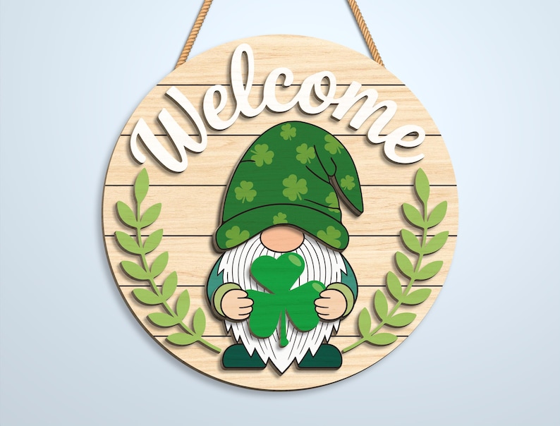 K&ouml;nnte beinhalten: Ein rundes h&auml;ngendes Holzschild mit dem Wort Welcome in wei&szlig;er Schreibschrift. In der Mitte befindet sich ein Gnom mit einem gr&uuml;nen Hut mit Kleeblattmuster, der ein gro&szlig;es gr&uuml;nes Kleeblatt h&auml;lt. Zwei hellgr&uuml;ne Blattzweige umrahmen den Gnom.