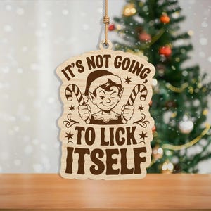 May include: Wooden Christmas ornament with a humorous elf design. The ornament features an elf winking and holding candy canes, with the phrase "It's not going to lick itself." The ornament is hanging in front of a decorated Christmas tree.