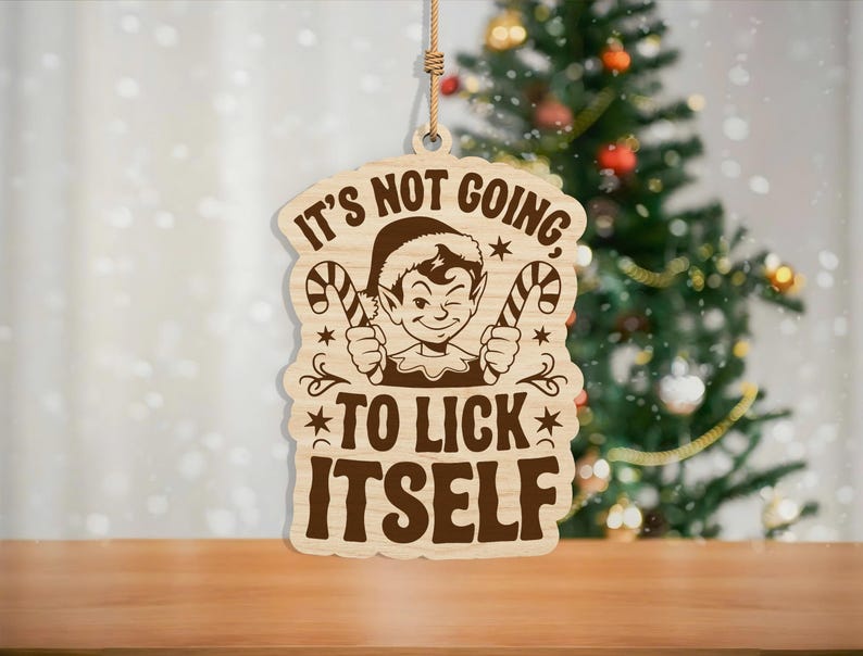 May include: Wooden Christmas ornament with a humorous elf design. The ornament features an elf winking and holding candy canes, with the phrase "It's not going to lick itself." The ornament is hanging in front of a decorated Christmas tree.