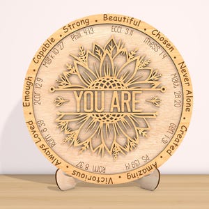 May include: A wooden round plaque with a sunflower design and the words "YOU ARE" in the center. The plaque is surrounded by a circle of words and bible verses, including "Strong", "Beautiful", "Chosen", "Never Alone", "Created", "Amazing", "Victorious", "Always Loved", "Enough", "Capable".