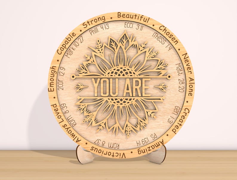 May include: A wooden round plaque with a sunflower design and the words "YOU ARE" in the center. The plaque is surrounded by a circle of words and bible verses, including "Strong", "Beautiful", "Chosen", "Never Alone", "Created", "Amazing", "Victorious", "Always Loved", "Enough", "Capable".