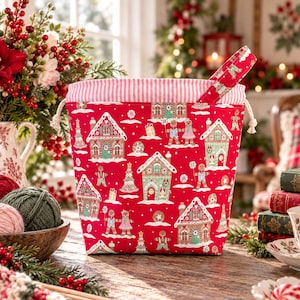 May include: A red fabric drawstring bag featuring a repeating pattern of gingerbread houses, gingerbread people, and Christmas trees. The bag has a white and red striped interior lining and a matching fabric handle attached to the top edge.