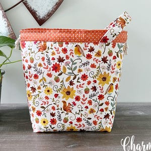 May include: A fabric storage basket featuring a white background with a colorful autumn-themed print of squirrels, birds, sunflowers, mushrooms, and leaves. The top edge has an orange band with white polka dots and a drawstring closure.