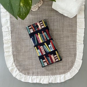 May include: A rectangular fabric case featuring a black background with a repeating pattern of colorful book spines. The books display various titles including Heidi, Gulliver's Travels, and Treasure Island. The case has a single black snap closure.