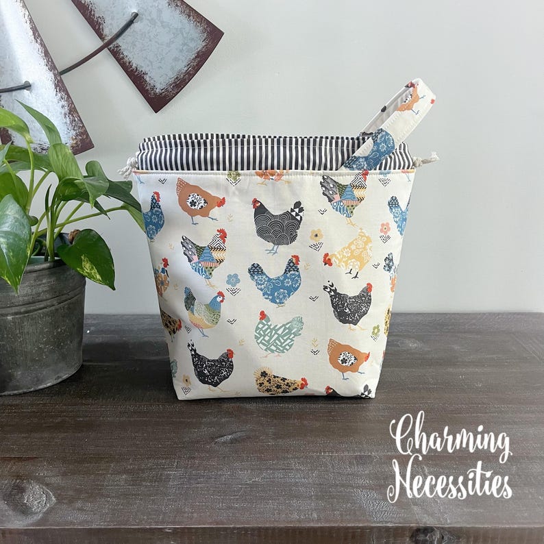 A charming chicken-themed project bag with a striped interior and a handle, placed on a wooden surface next to a potted plant.