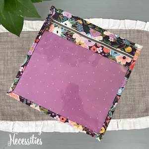 May include: A square zippered pouch featuring a dark fabric border with a colorful floral pattern of pink, orange, and blue flowers. The center of the pouch is made of a translucent purple material with a subtle pattern of small white dots.
