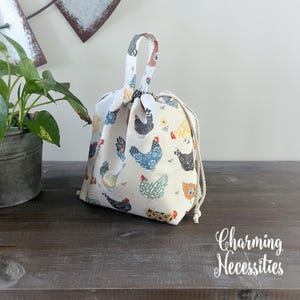 A white drawstring bag with a chicken print pattern is placed on a wooden surface next to a potted plant.