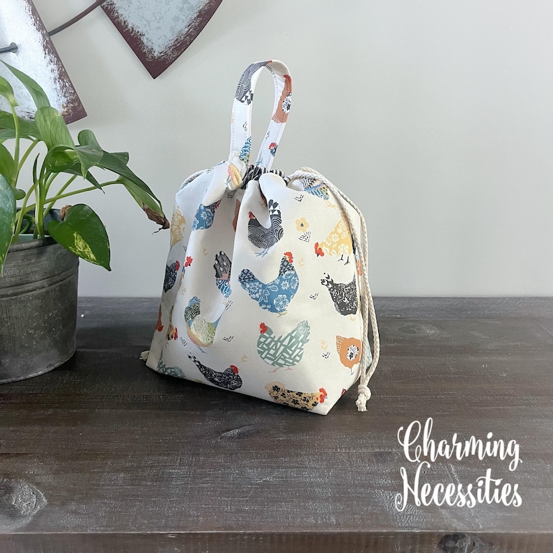 A white drawstring bag with a chicken print pattern is placed on a wooden surface next to a potted plant.