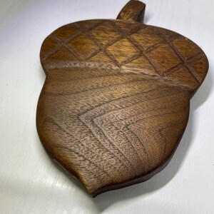 May include: A handcrafted wooden acorn-shaped trivet. The trivet is made of dark brown wood with a diamond pattern carved into the top portion. The trivet has a small handle and is designed to protect surfaces from hot items.