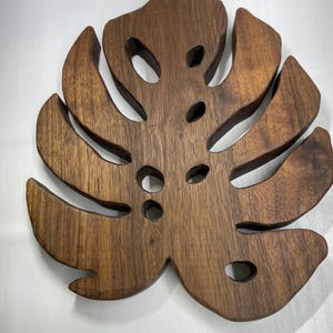 May include: A wooden coaster in the shape of a Monstera leaf. The coaster is made of dark brown wood and features several cut-out holes. The leaf design is detailed and has a natural wood grain appearance. The coaster is designed to protect surfaces from heat and moisture.