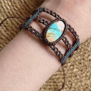 May include: A macrame bracelet featuring a central oval labradorite gemstone with iridescent blue and gold tones. The band is constructed from dark brown cord with intricate knotting, accented by small metallic beads and thin teal thread details.