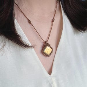 Könnte beinhalten: Eine Halskette mit einem quadratischen, goldfarbenen Edelsteinanhänger, der in braune Makramee-Schnur eingefasst ist. Die Schnur ist mit kleinen Metallperlen verziert und geht in eine dünne, braune geflochtene Kette über.