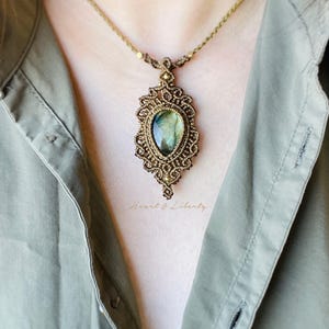 May include: A handmade macrame necklace with a large, oval gemstone pendant. The pendant has green and blue tones, set in an ornate, bronze-coloured macrame frame. The necklace is worn against a green shirt, with the text "Heart & Liberty".