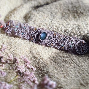 Pode incluir: Uma pulseira de macramé feita de cordão cor de malva com um padrão entrelaçado intrincado, uma pedra preciosa central de labradorite azul de forma oval e pequenas contas incrustadas em tons de turquesa, âmbar e vermelho escuro no design.