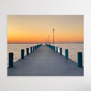 May include: A wooden pier extends out into a calm body of water at sunrise. The pier is lined with blue railings and has several streetlights along the way. The sky is a soft orange and pink.