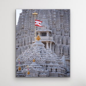 May include: A white marble temple with intricate carvings and a red flag flying from a gold pole. The temple has multiple domes and towers, and the flag is visible against the clear sky.