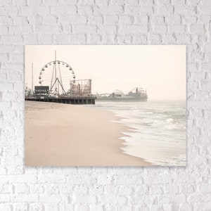 May include: A sepia-toned photograph of a beach with an amusement park pier. A Ferris wheel and roller coaster are visible on the pier. The beach has light-colored sand, and the ocean has gentle waves. The sky is overcast.